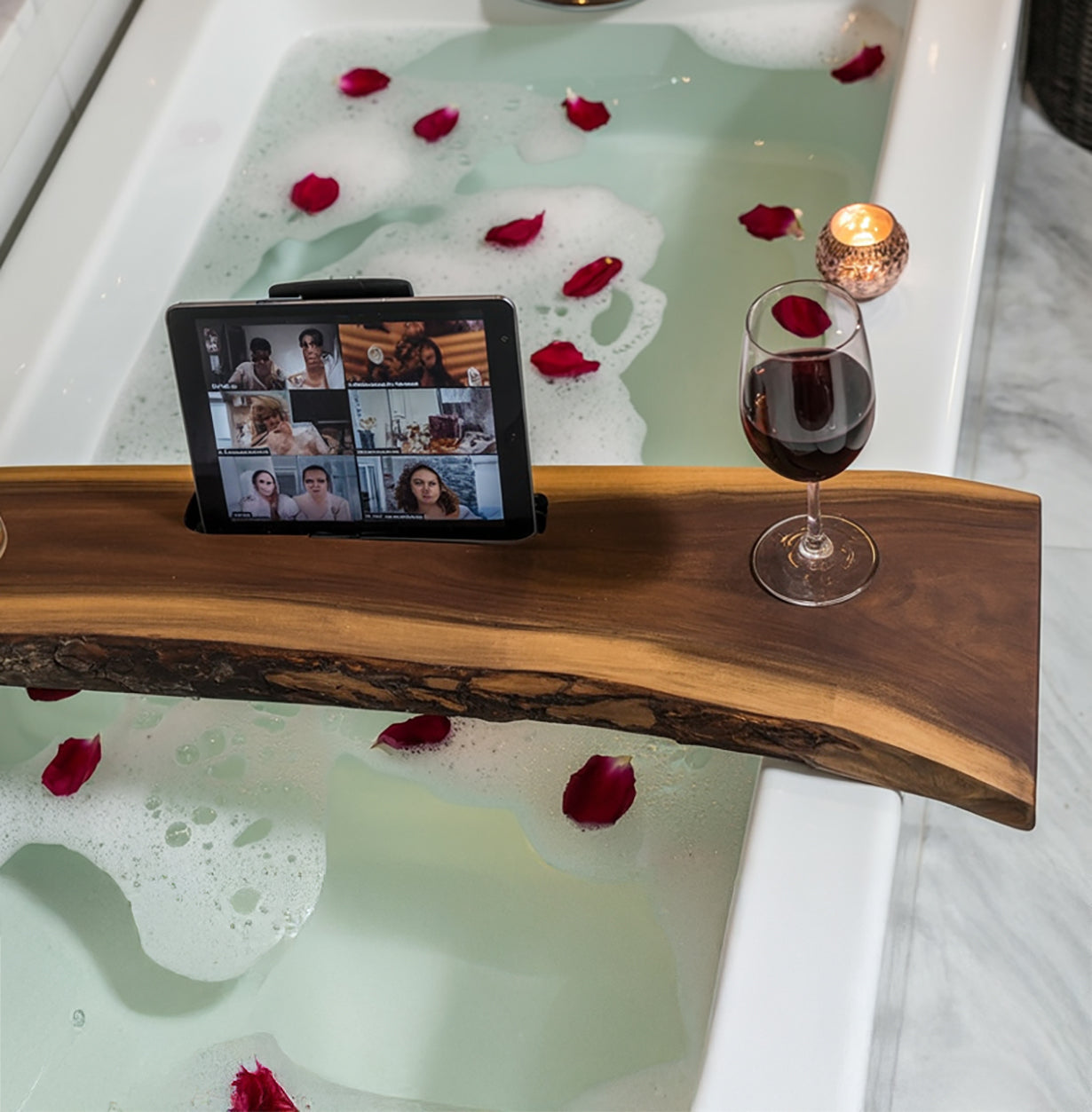 Wooden bathtub tray with tablet, wine glass, candle, and rose petals on foam-filled bath