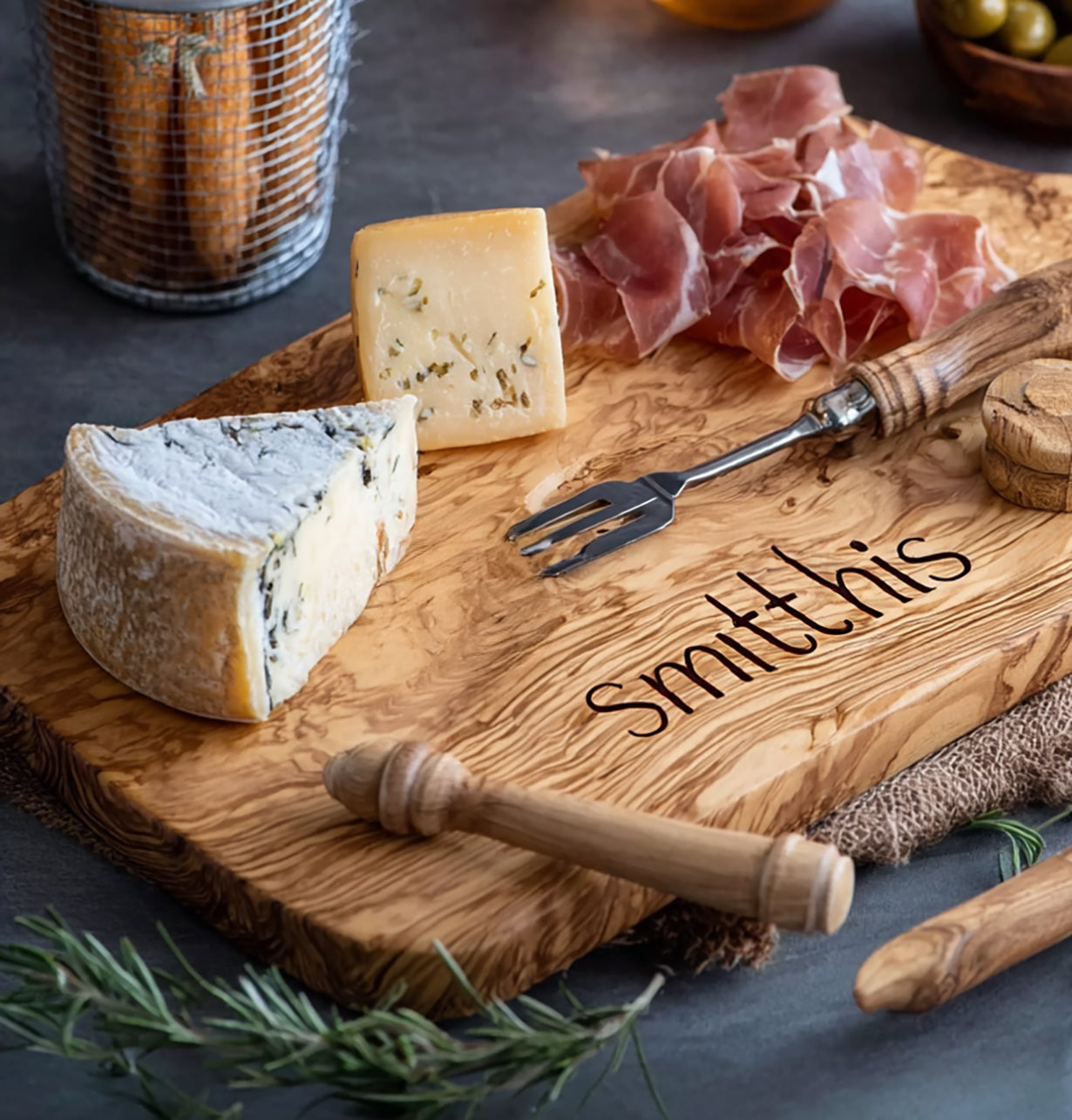 Wooden cheese board with the name Smith this, blue cheese wedge, herb cheese, prosciutto slices, small fork, and rosemary sprig