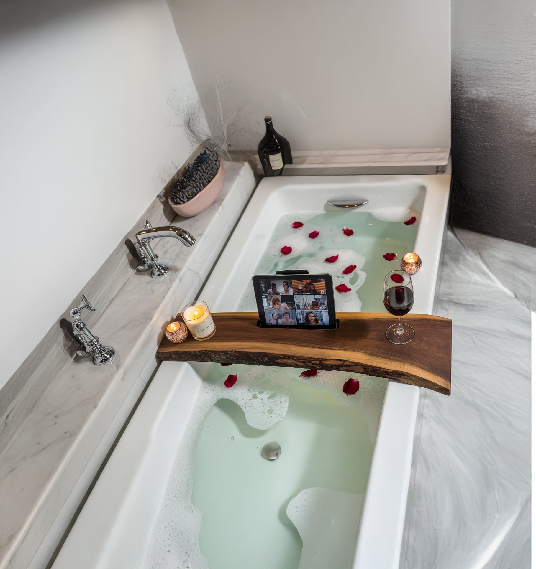 White bathtub with rose petals, wooden bath tray holding candles, tablet, and glass of red wine