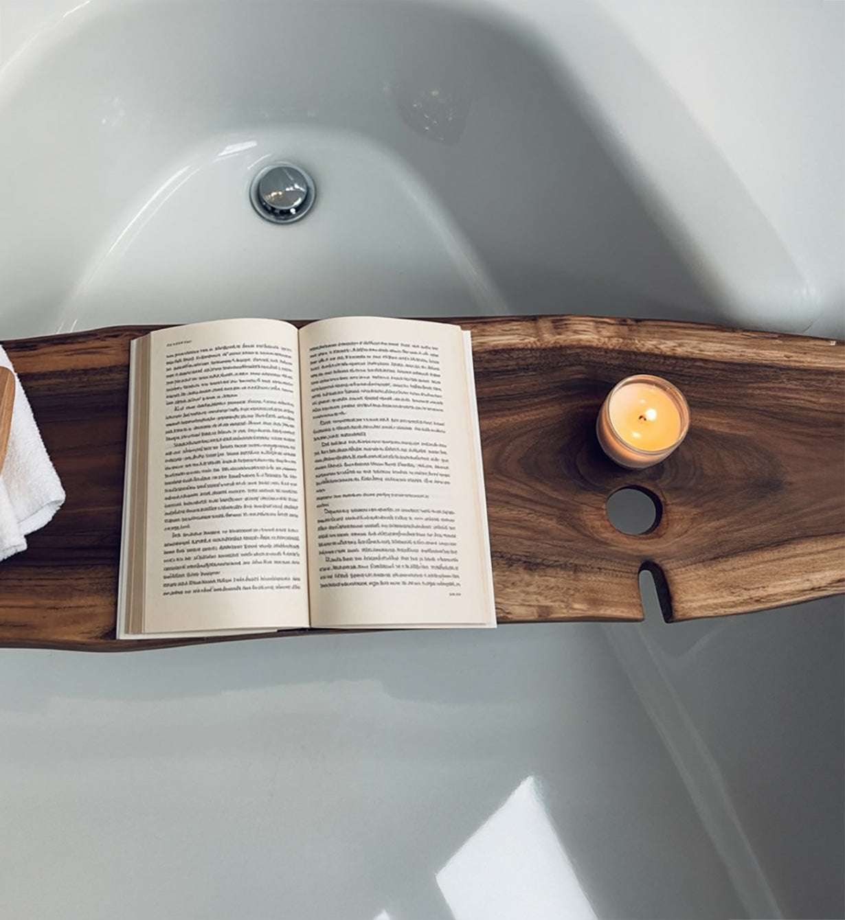 Wooden bathtub tray holding an open book and a lit candle over a white bathtub
