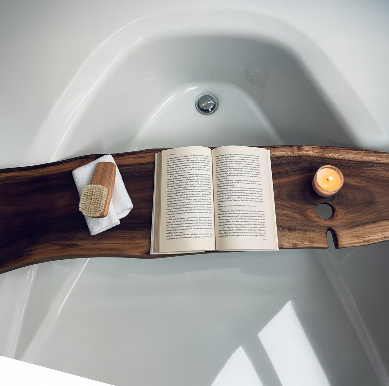 Wooden bathtub caddy holding an open book, candle, towel, and brush on a white bathtub