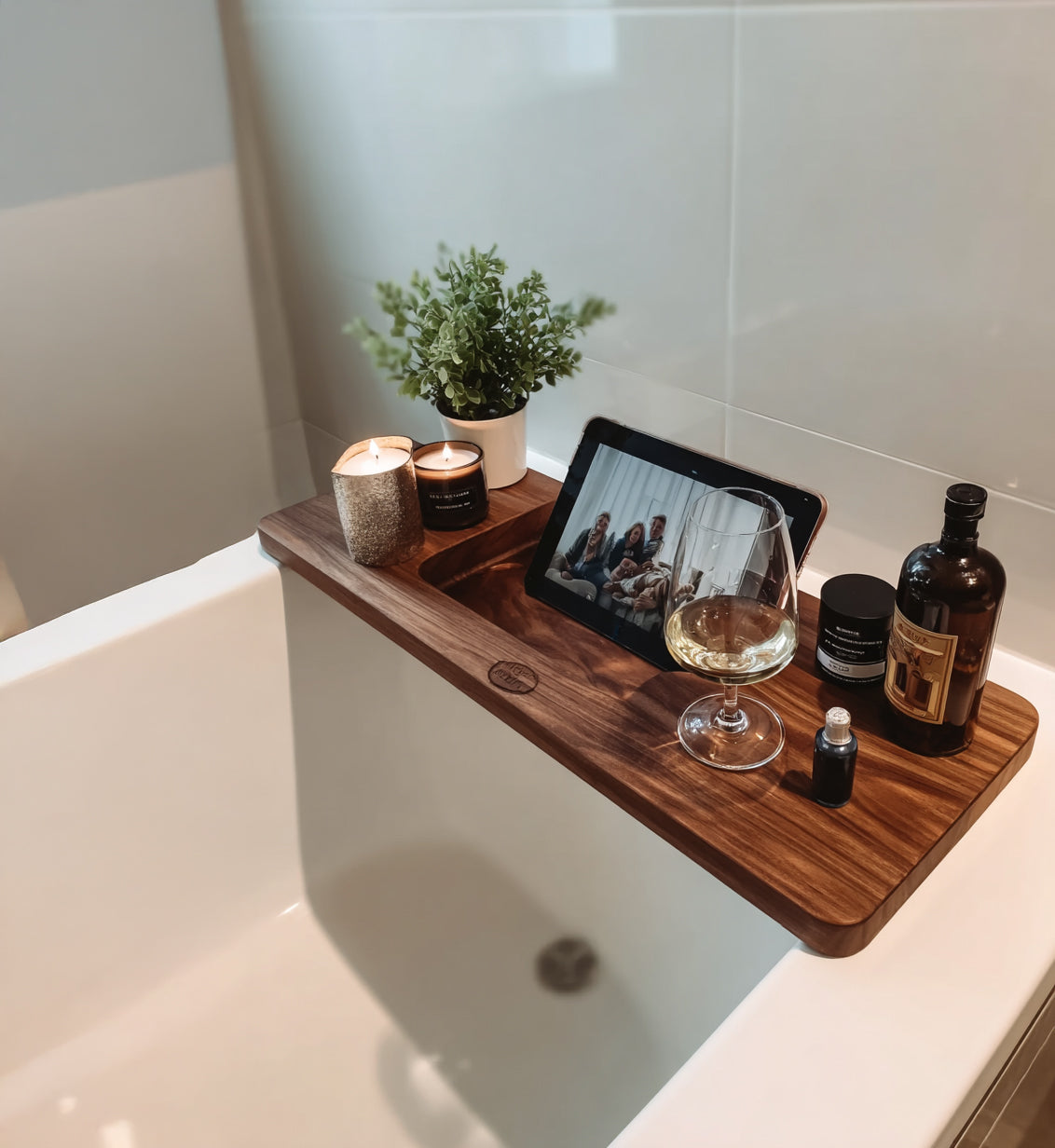 Wooden bathtub tray holding lit candles, small plant, wine glass, tablet showing video call, and bath products