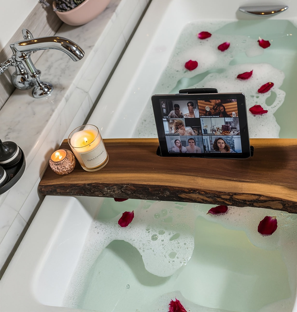 Wooden bath tray with lit candles and tablet showing video call over bathtub with rose petals and foam