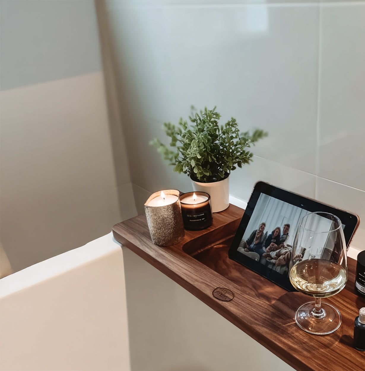 Wooden bathtub tray with lit candles, potted plant, tablet, and glass of white wine in bathroom