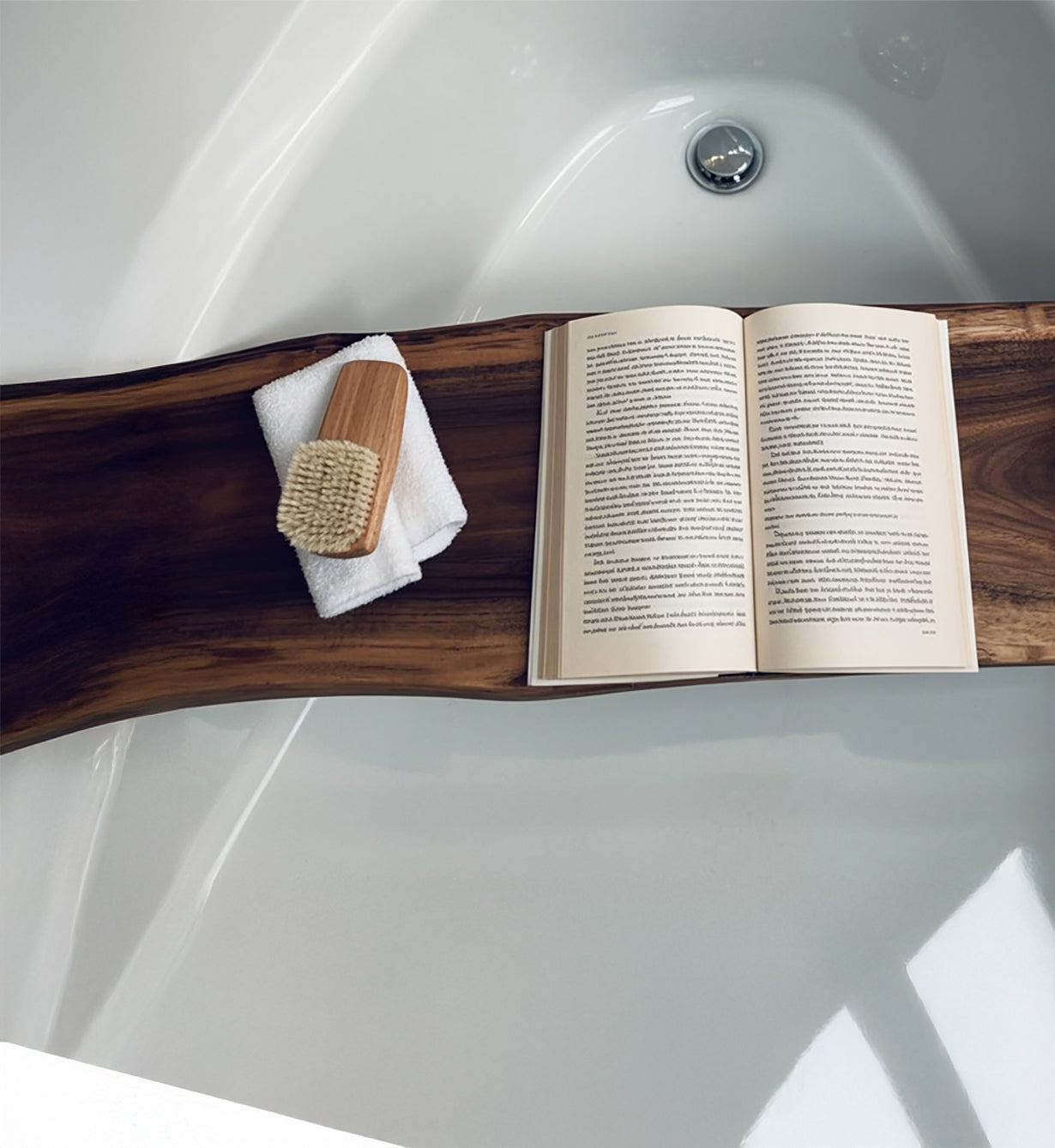 Wooden bath tray over white bathtub holding an open book, white towel, and wooden brush
