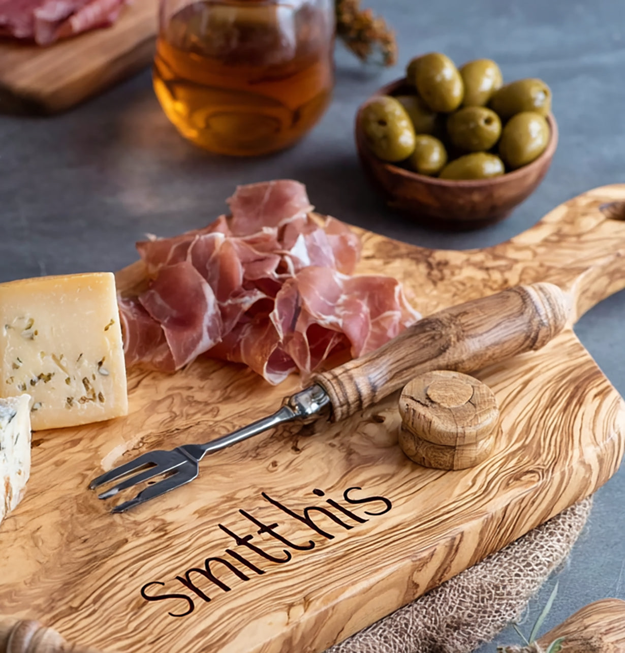 Wooden charcuterie board engraved with name, holding prosciutto, cheese, olives, and a serving fork