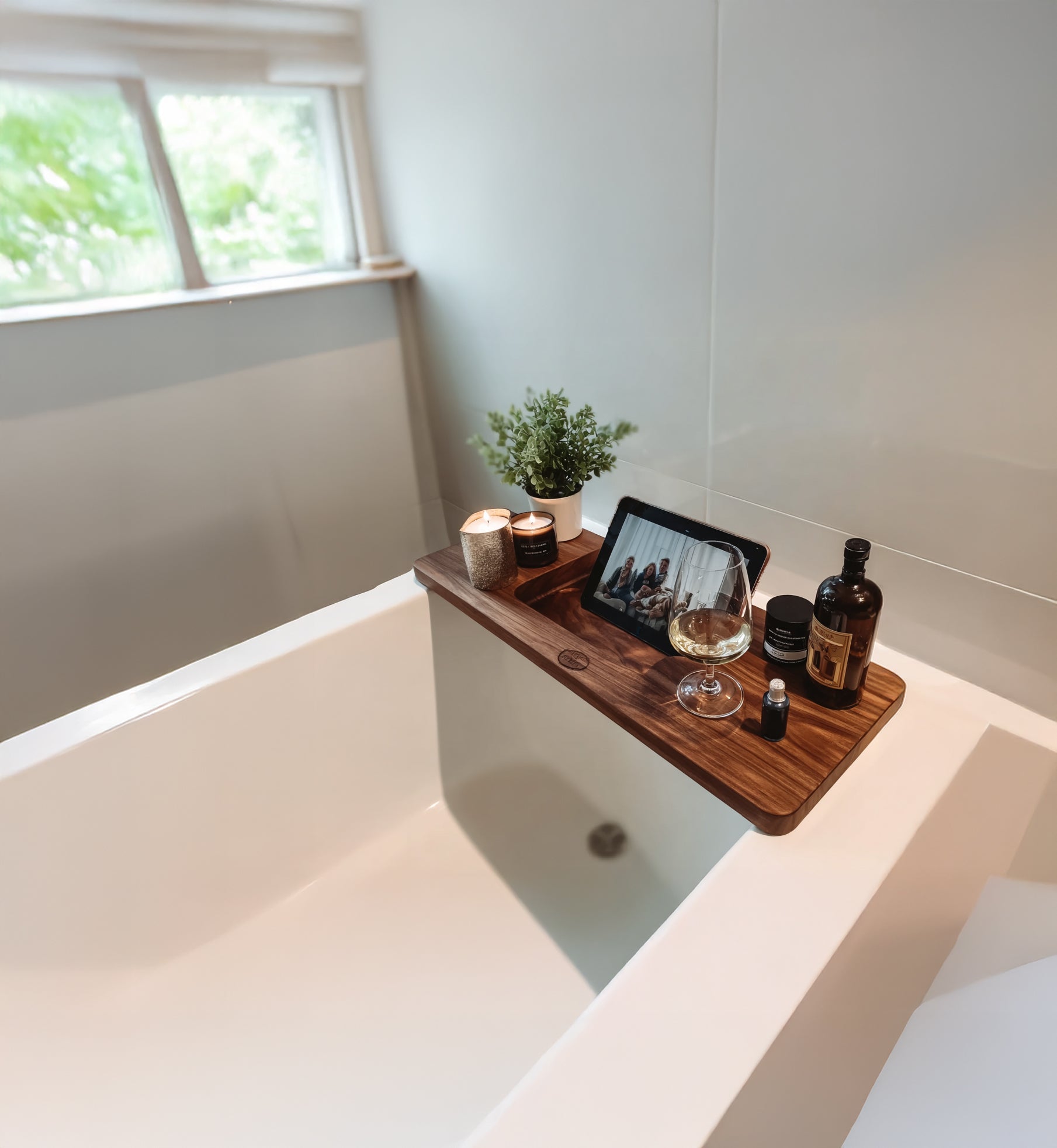 Modern white bathtub with wooden tray holding candles, plant, wine glass, bottle, and tablet showing a group photo