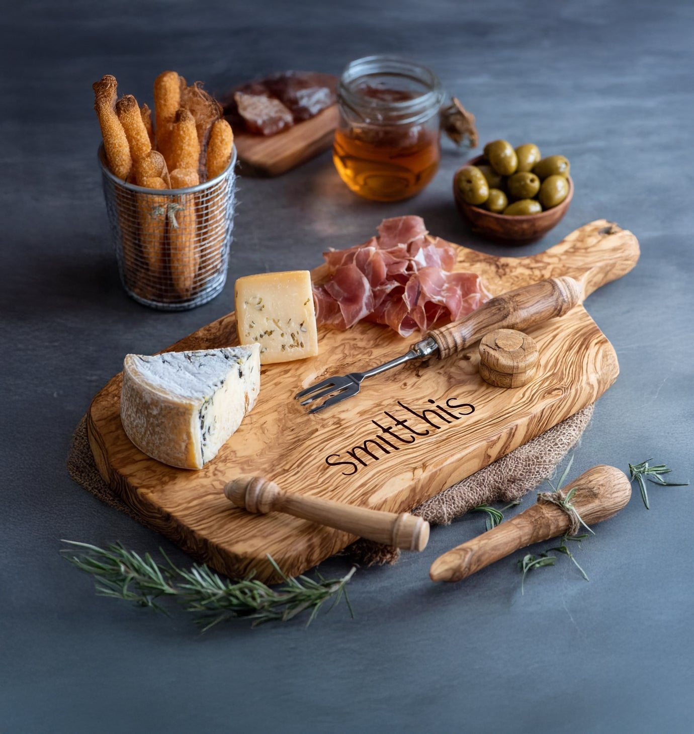 Wooden charcuterie board with assorted cheeses, prosciutto, olives, breadsticks, honey, and rosemary
