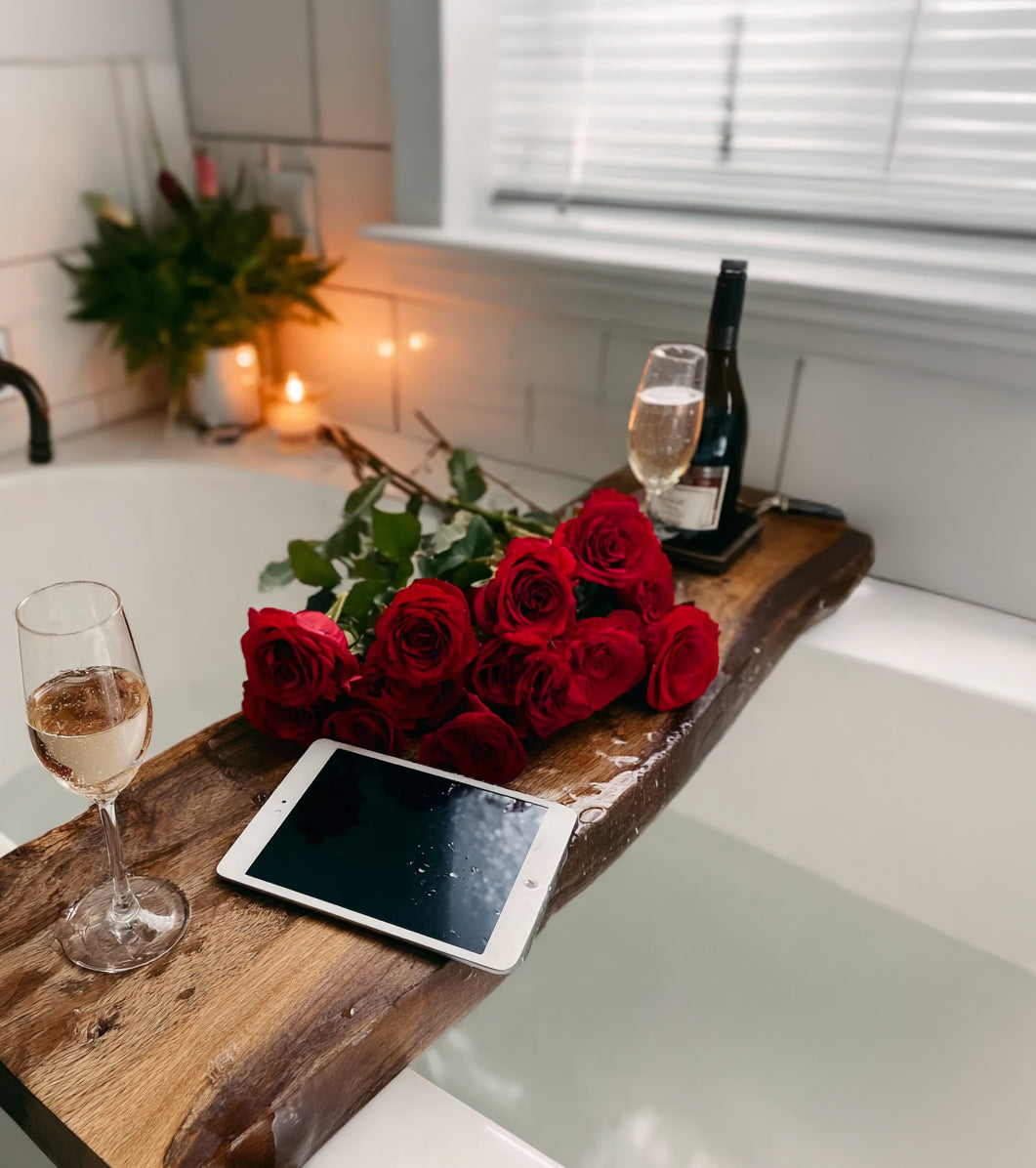 Wooden bath tray with red roses, tablet, wine bottle, and two glasses beside a lit candle in white bathroom