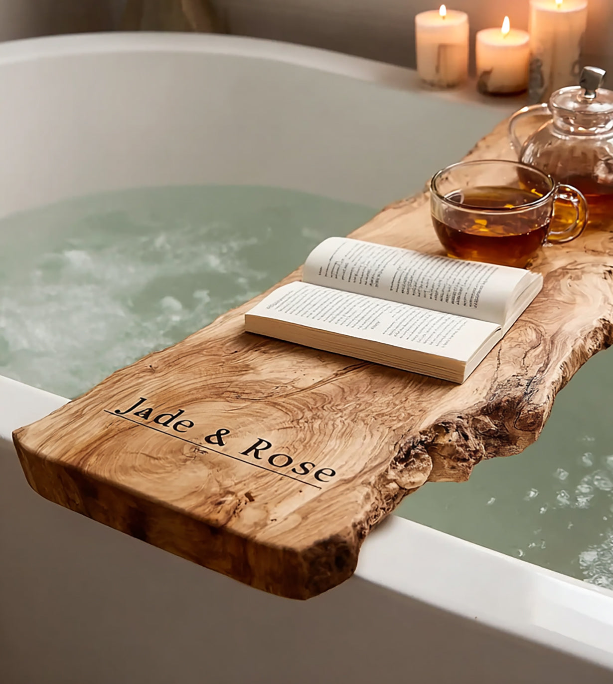 Wooden bath tray engraved with Jade & Rose holding a book and glass teapot cup over filled bathtub with lit candles
