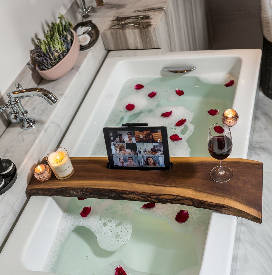 Wooden bath tray with lit candles, wine glass, and tablet over bathtub with rose petals and foam